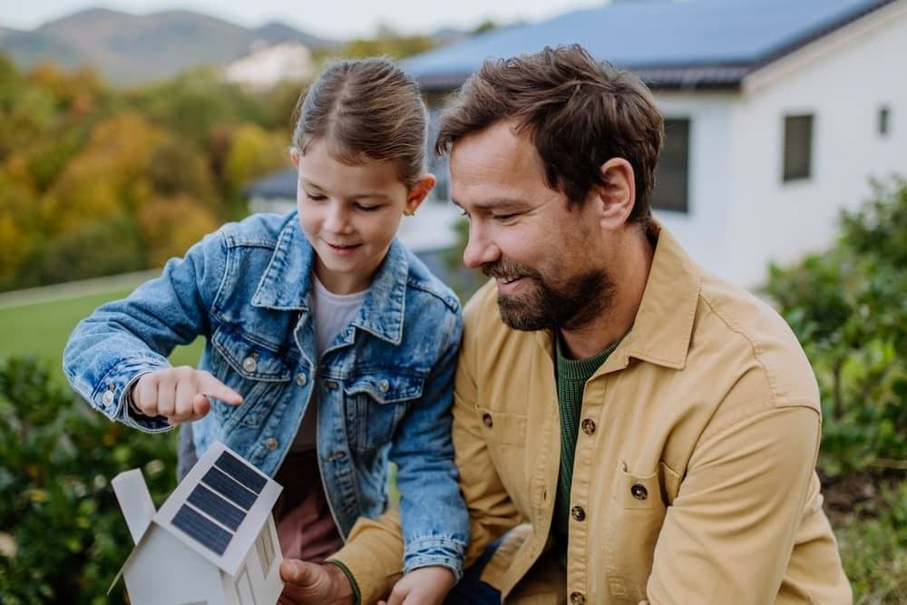 Energy Banner - Father looks at a toy house with solar panels with his daughter in front of their home with solar panels