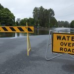 Water across road sign, in front of water covering a wide road.