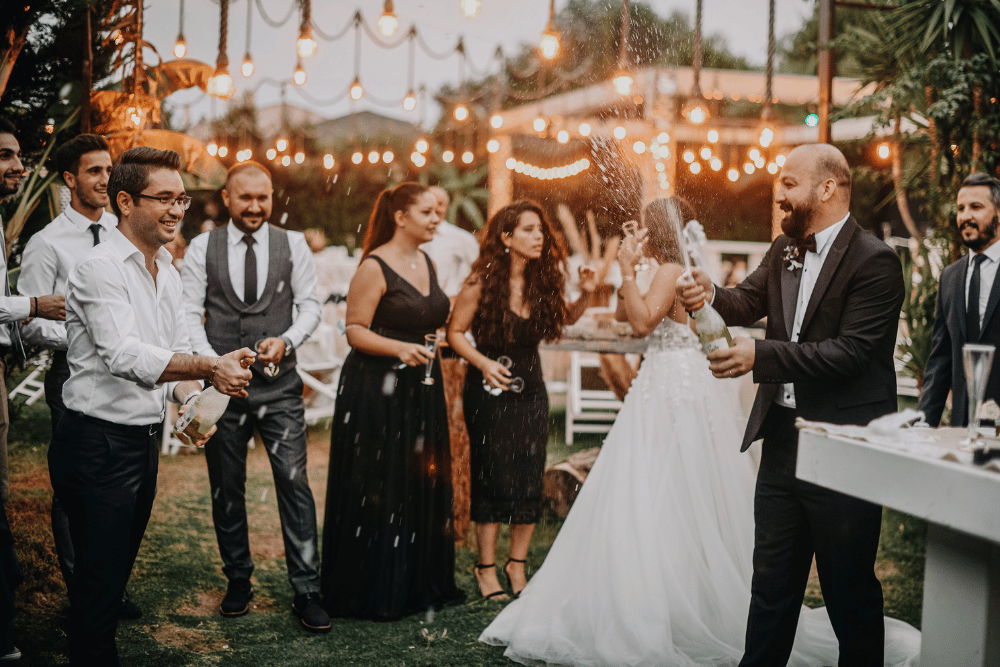 Groom opening a bottle of champagne at his wedding while his wife speaks to guests