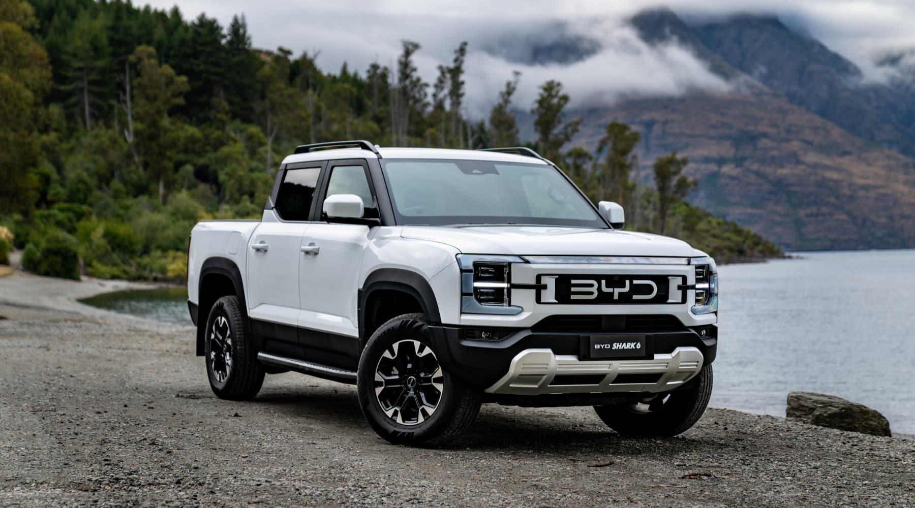 White BYD Shark6 parked on gravel next to a lake with trees in the background