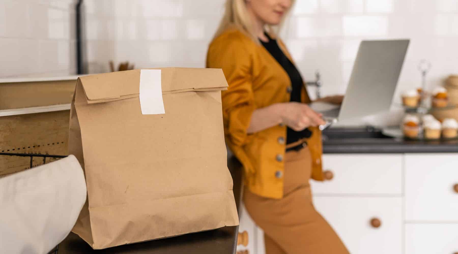 Woman standing in kitchen with laptop with takeaway food on counter